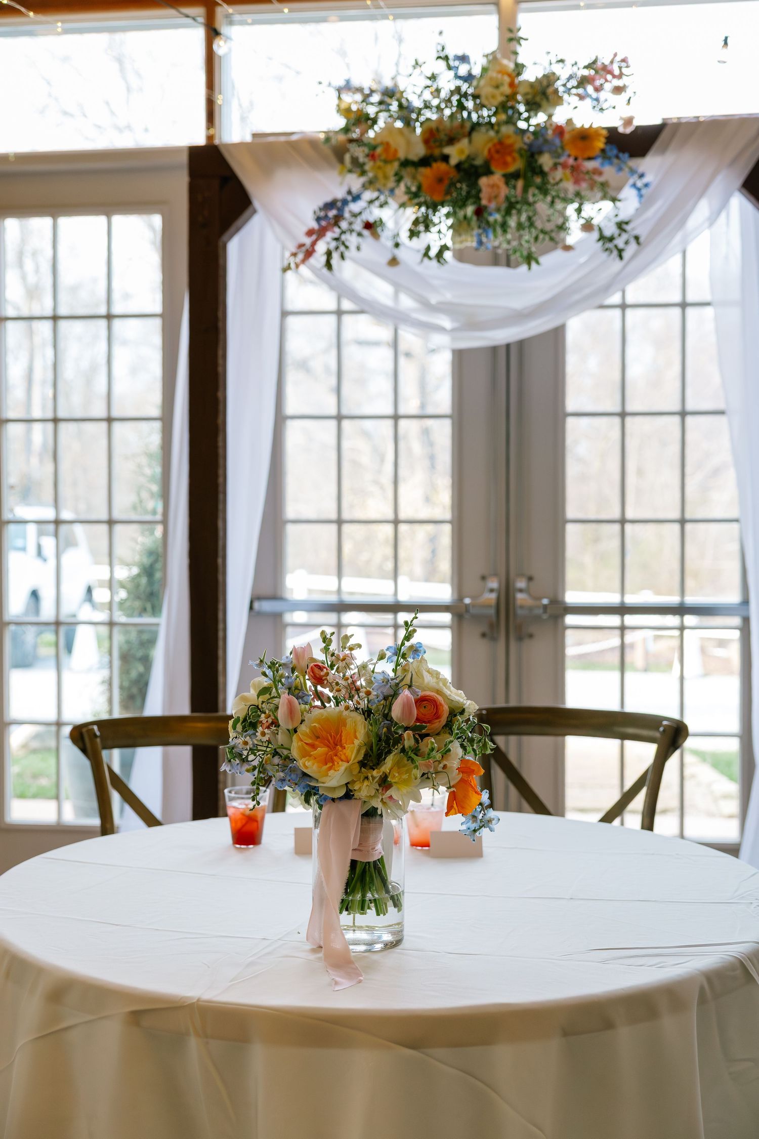 Romantic wedding sweetheart table with colorful bridal bouquet and floral chandelier backdrop near large windows.