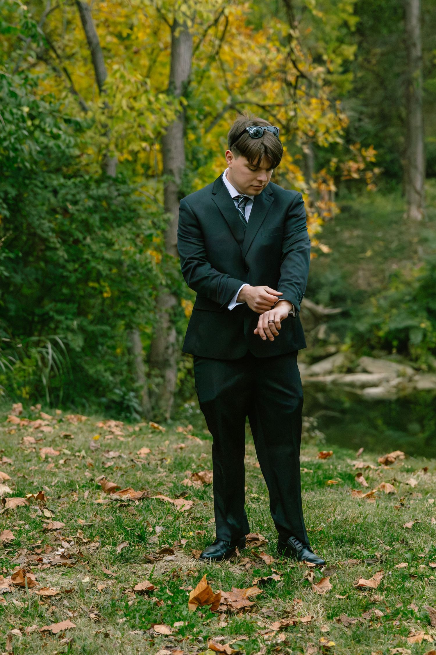 Dapper groom in black suit adjusts cufflinks while standing on grass surrounded by fall foliage.