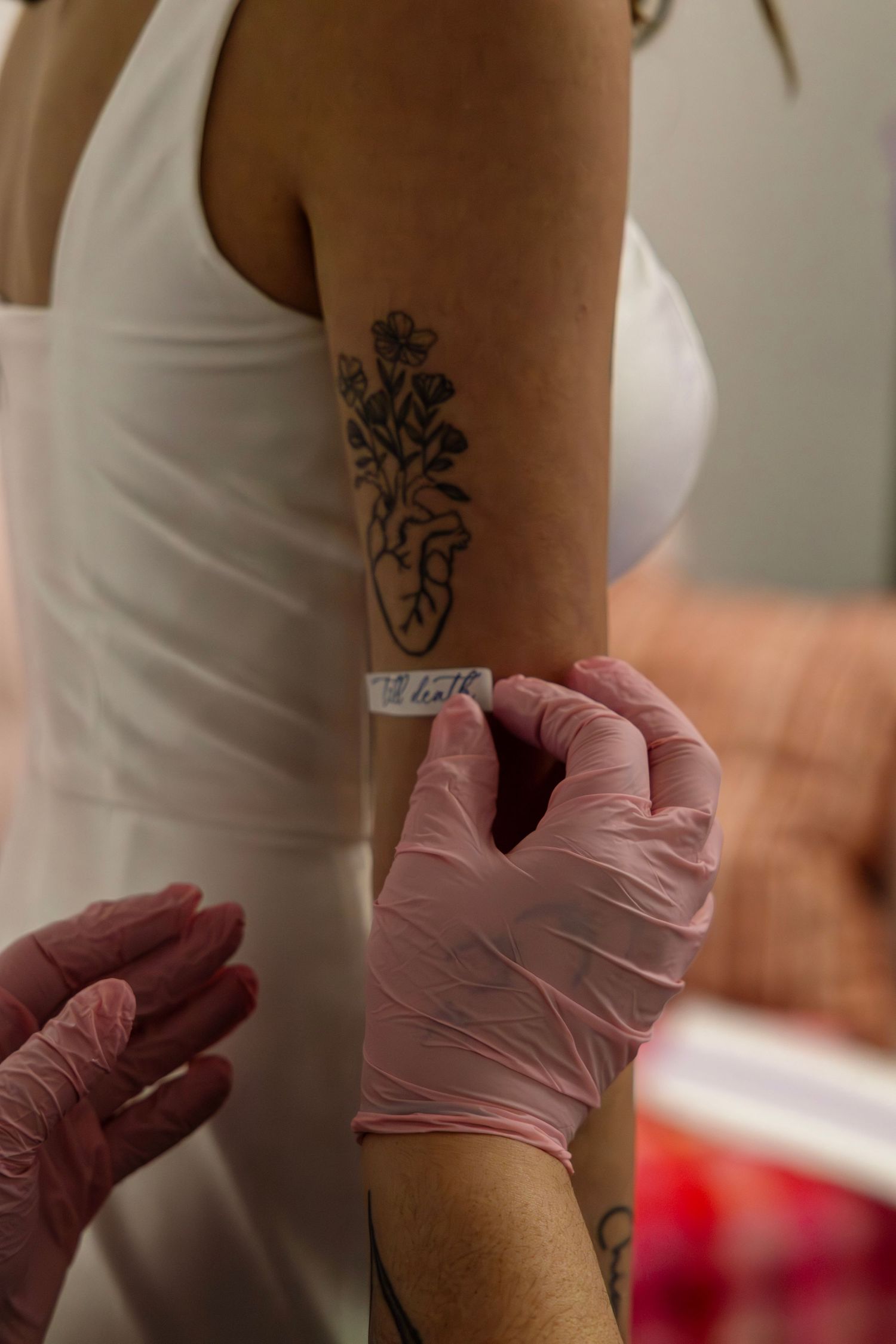 Tattoo artist in pink gloves applies stencil to bride's arm near heart and floral tattoo design.