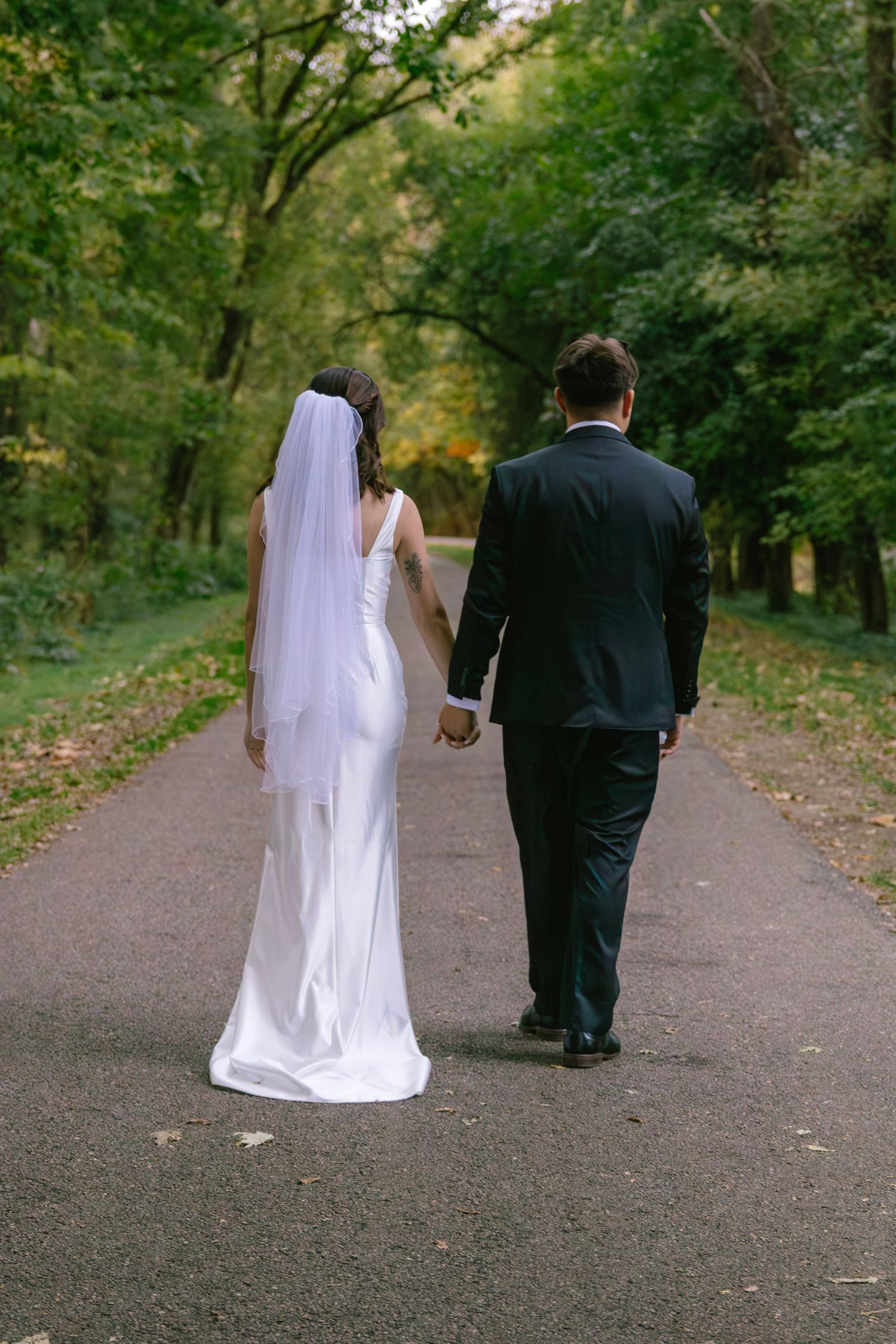 Bride in white gown and veil holds groom's hand walking away down scenic tree-lined park path.