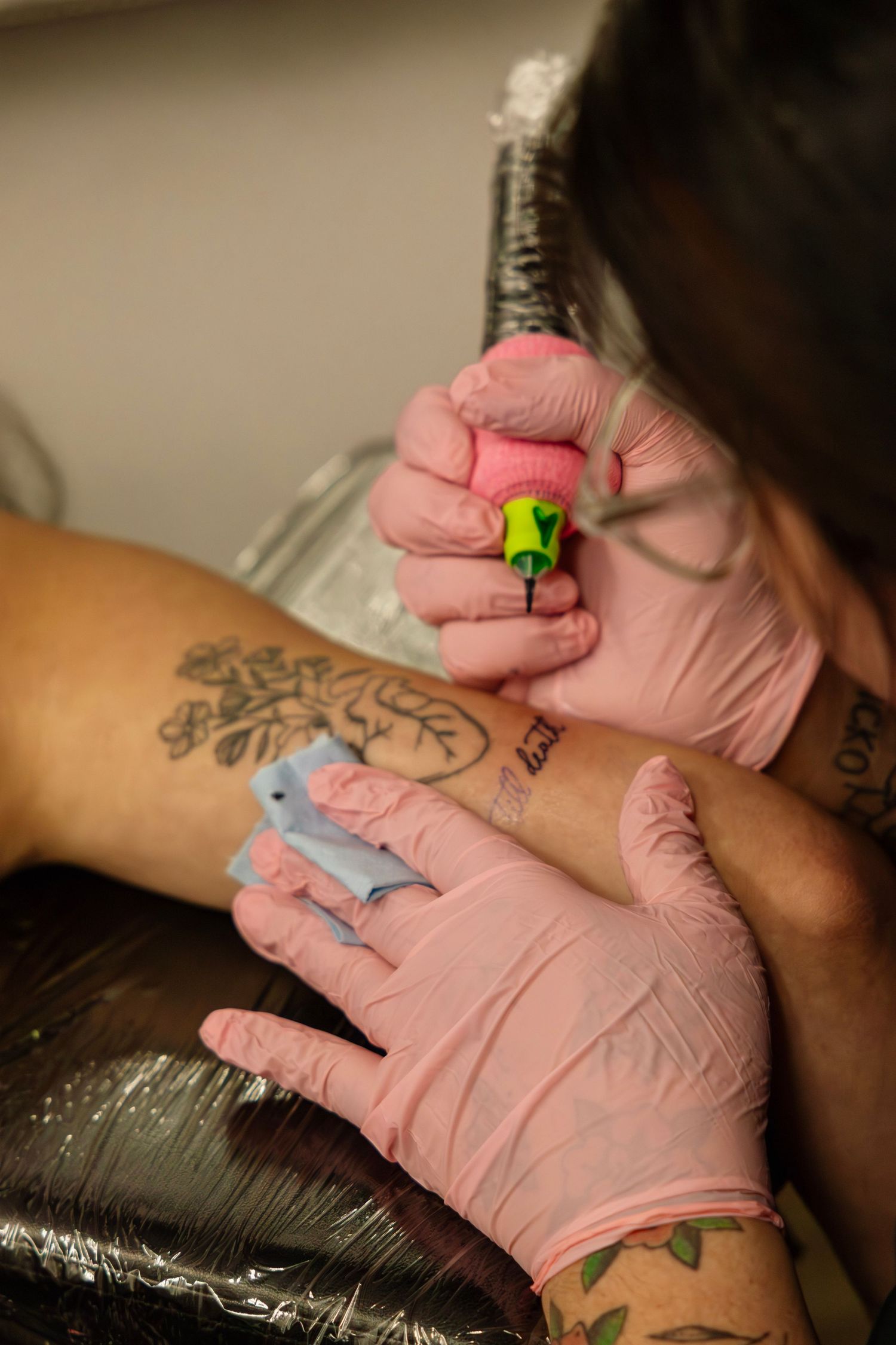 Close-up of tattoo artist in pink gloves tattooing bride's wrist near existing floral tattoo artwork.