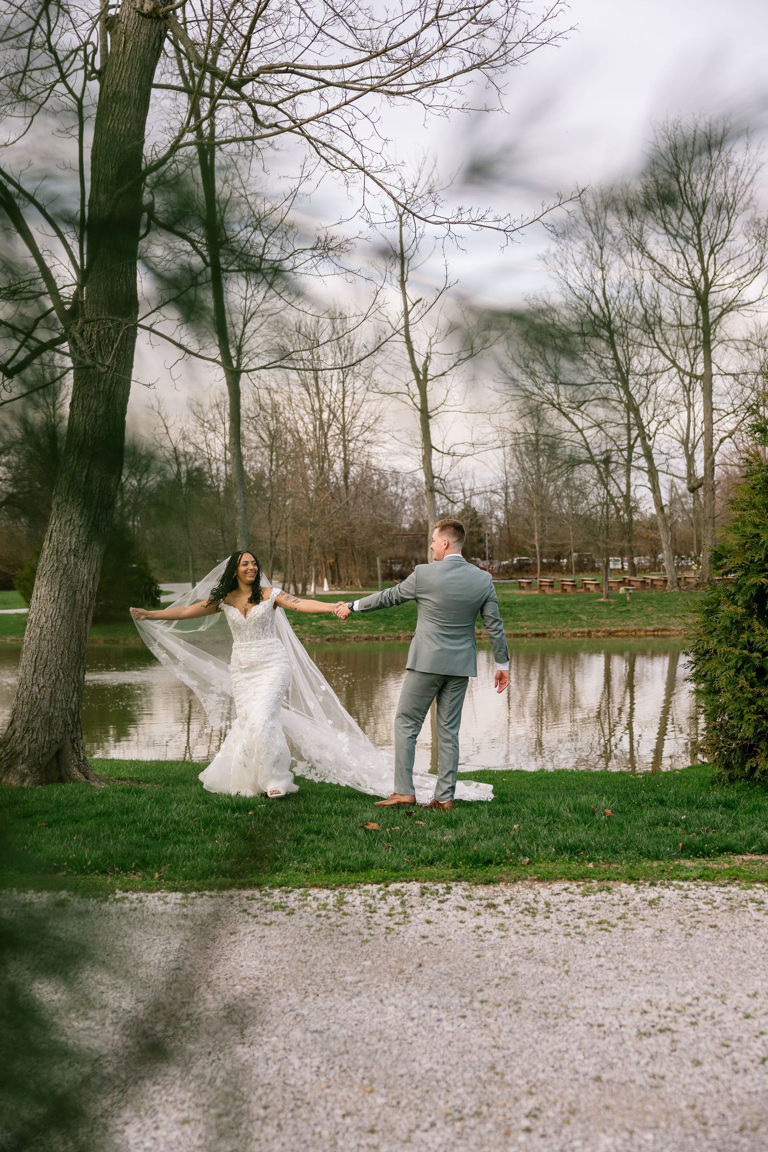 Bride and groom hold hands near a serene pond surrounded by bare trees during an outdoor wedding photo session.