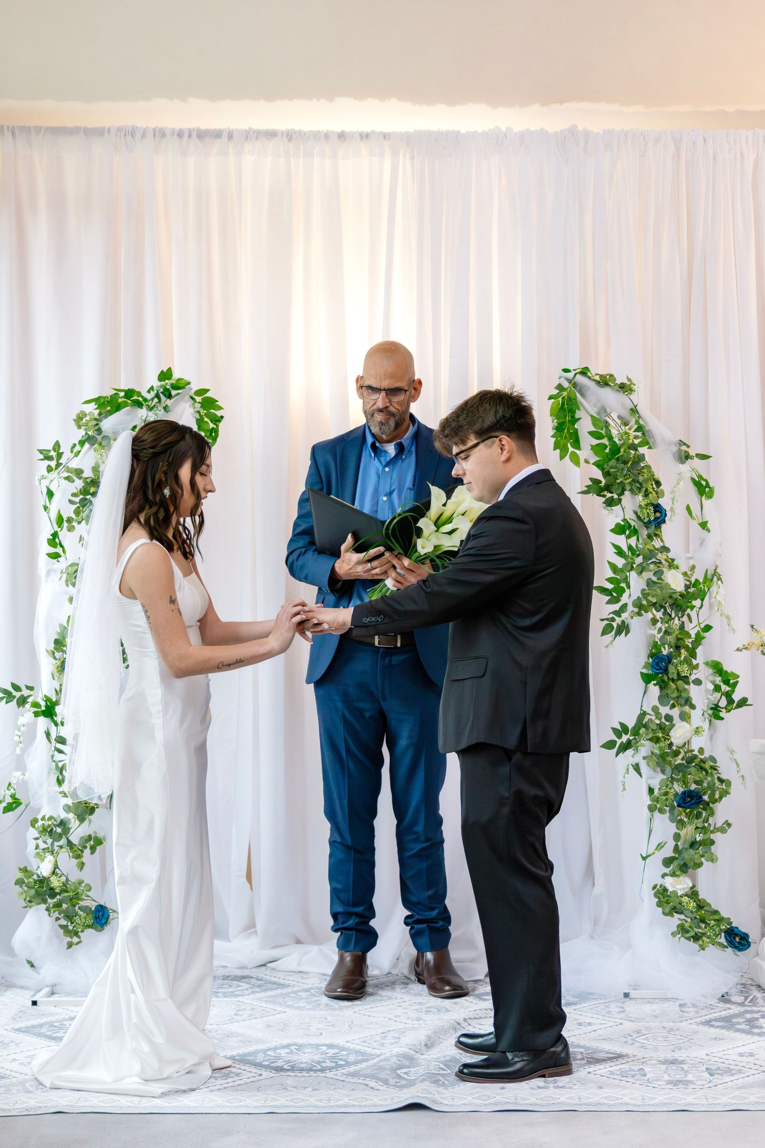 Couple exchanges rings at altar during wedding ceremony with officiant and white backdrop adorned with green ivy.