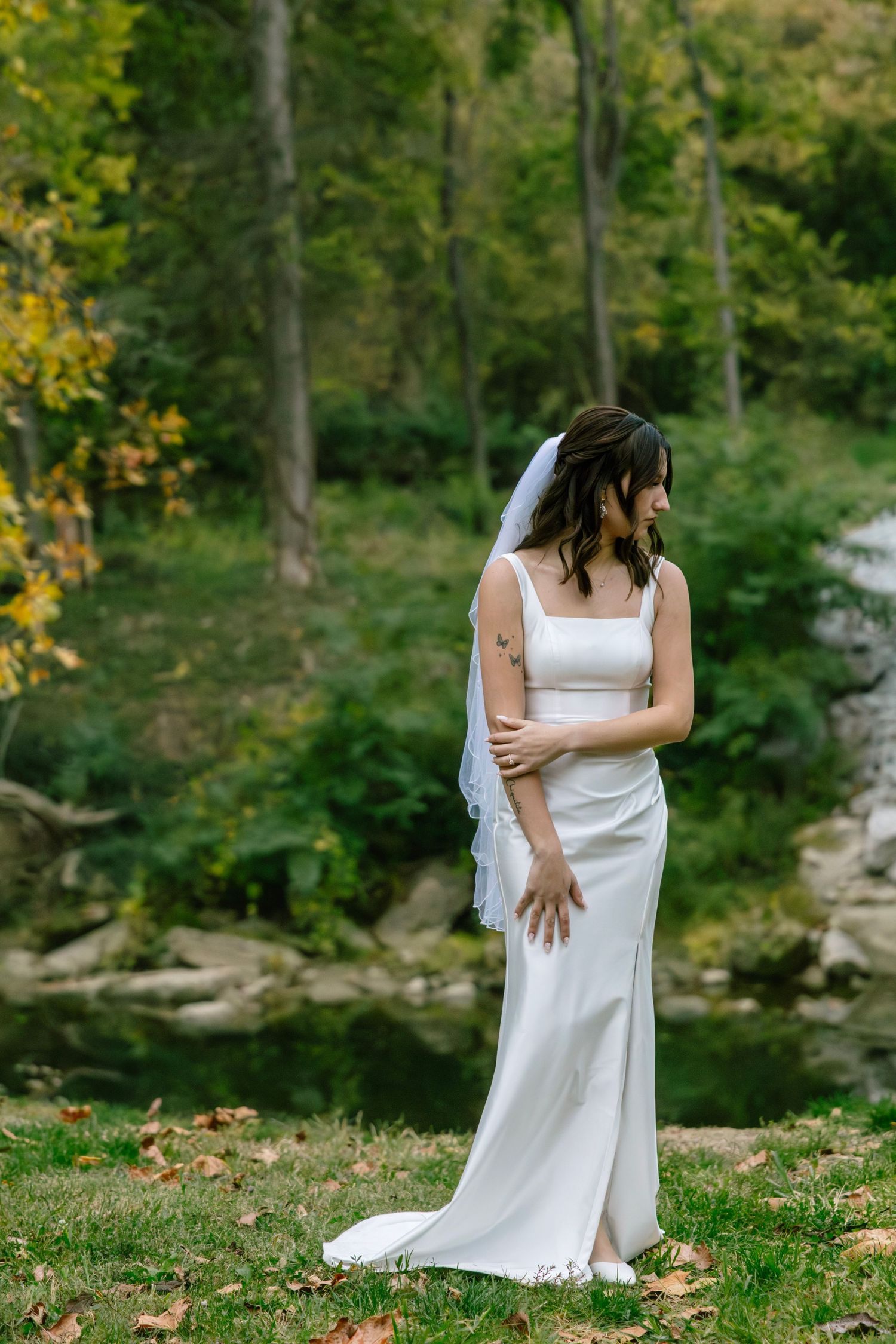 Elegant bride in fitted white satin gown and veil stands by peaceful pond in autumn forest.