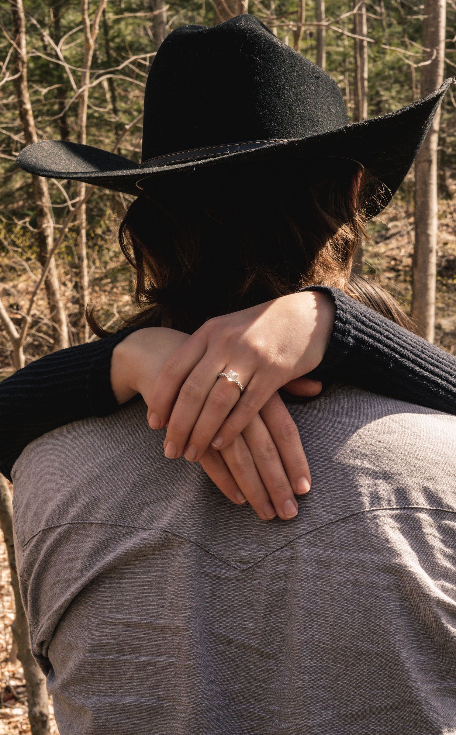Close-up of woman in black cowboy hat showing engagement ring while embracing partner outdoors.