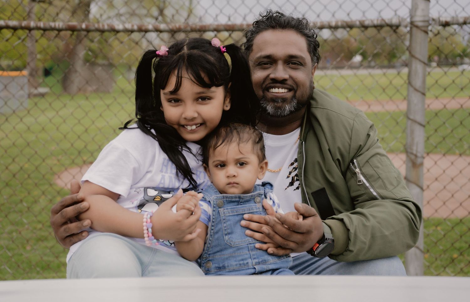 A man smiles with two young children outdoors, posing for a family photo in a park setting.