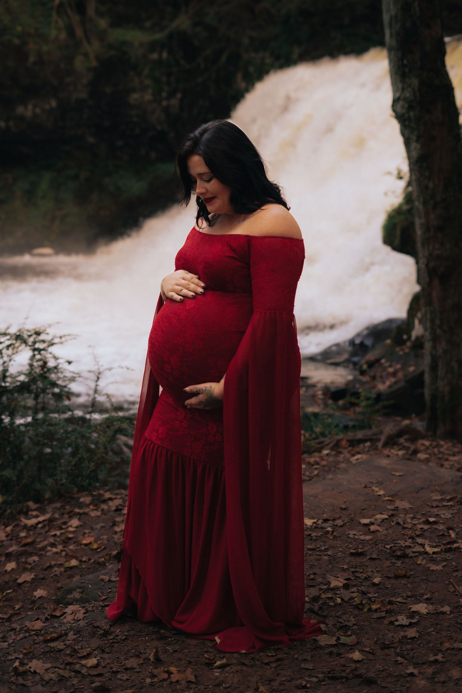 Pregnant woman in elegant red off-shoulder gown cradling belly near waterfall in forest.