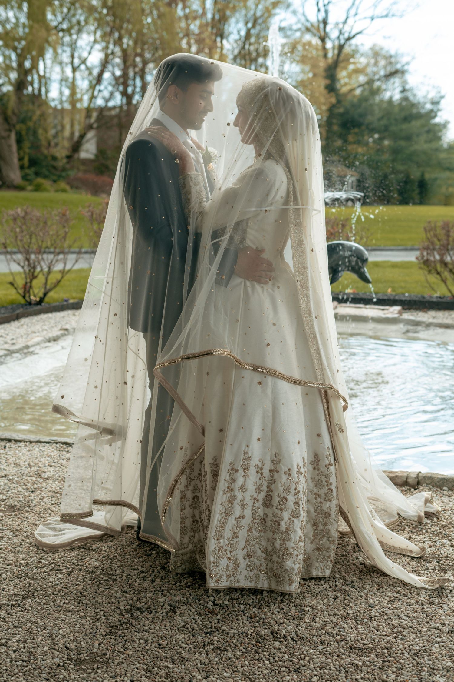 Bride and groom standing beneath a sheer veil near a fountain in a lush outdoor garden setting.