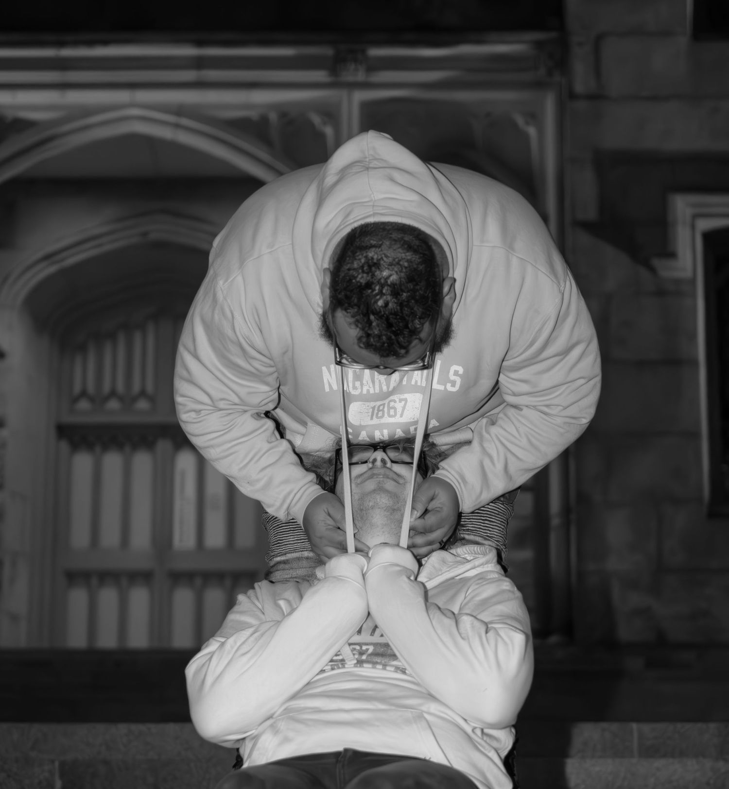 Man in hoodie bowing head in contemplative pose outside historic stone building in black and white.
