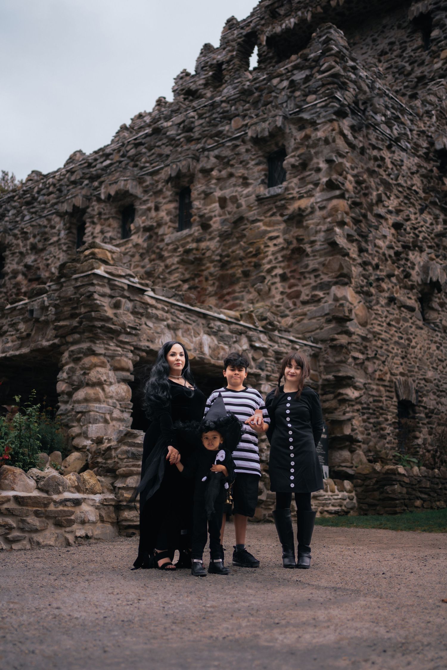 Family dressed in Addams Family Halloween costumes posing before ancient stone castle ruins.