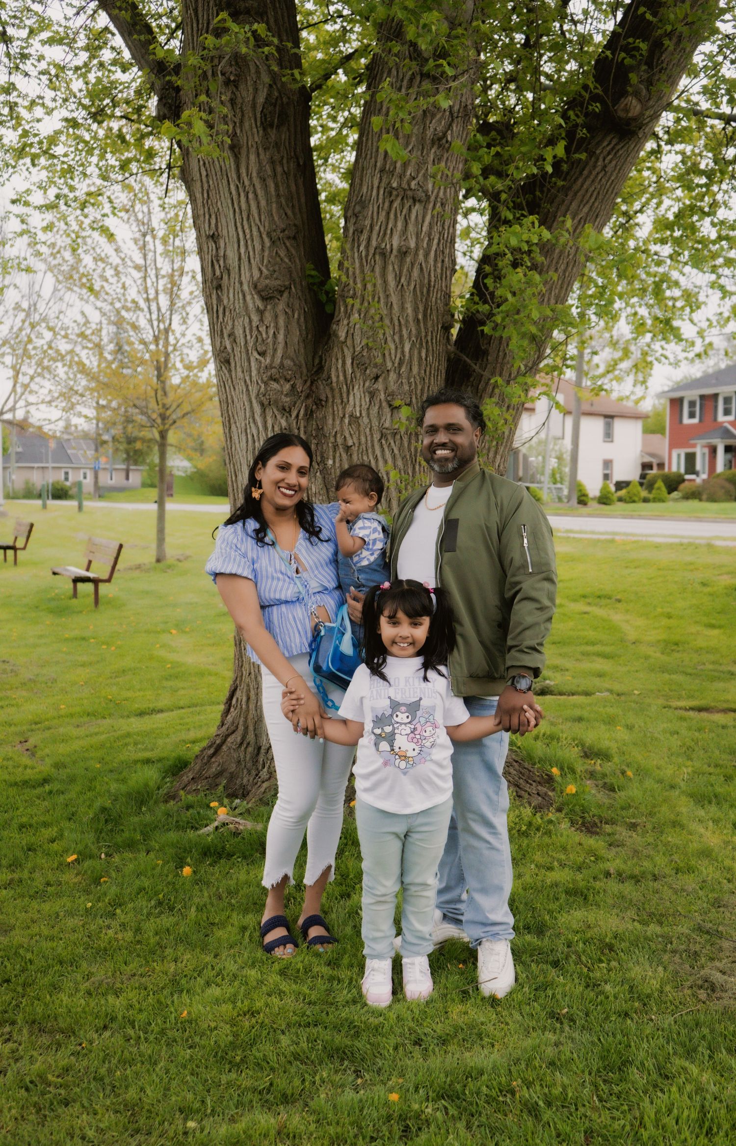 Family of four posing happily in front of a large tree in a green park on a spring day.