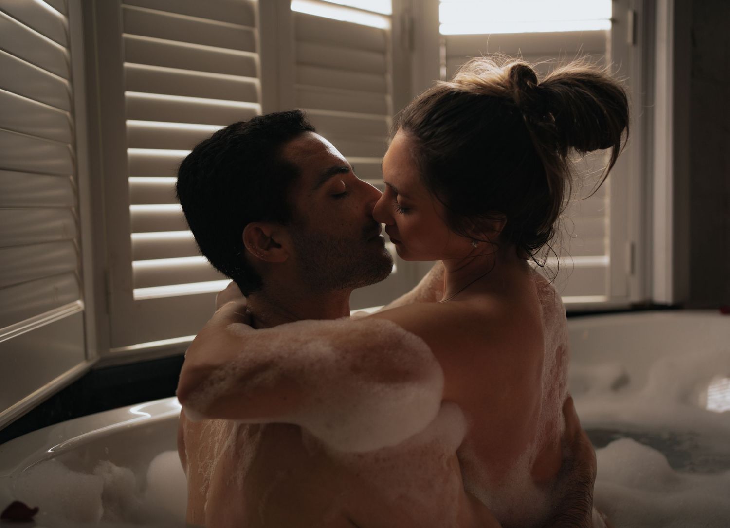 Couple sharing a romantic kiss in a bubble bath near shuttered windows.