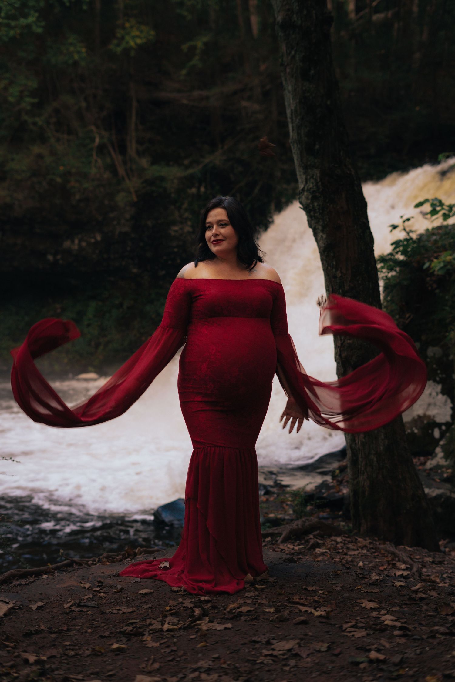 Pregnant woman in flowing red maternity gown with billowing sleeves posing by waterfall.