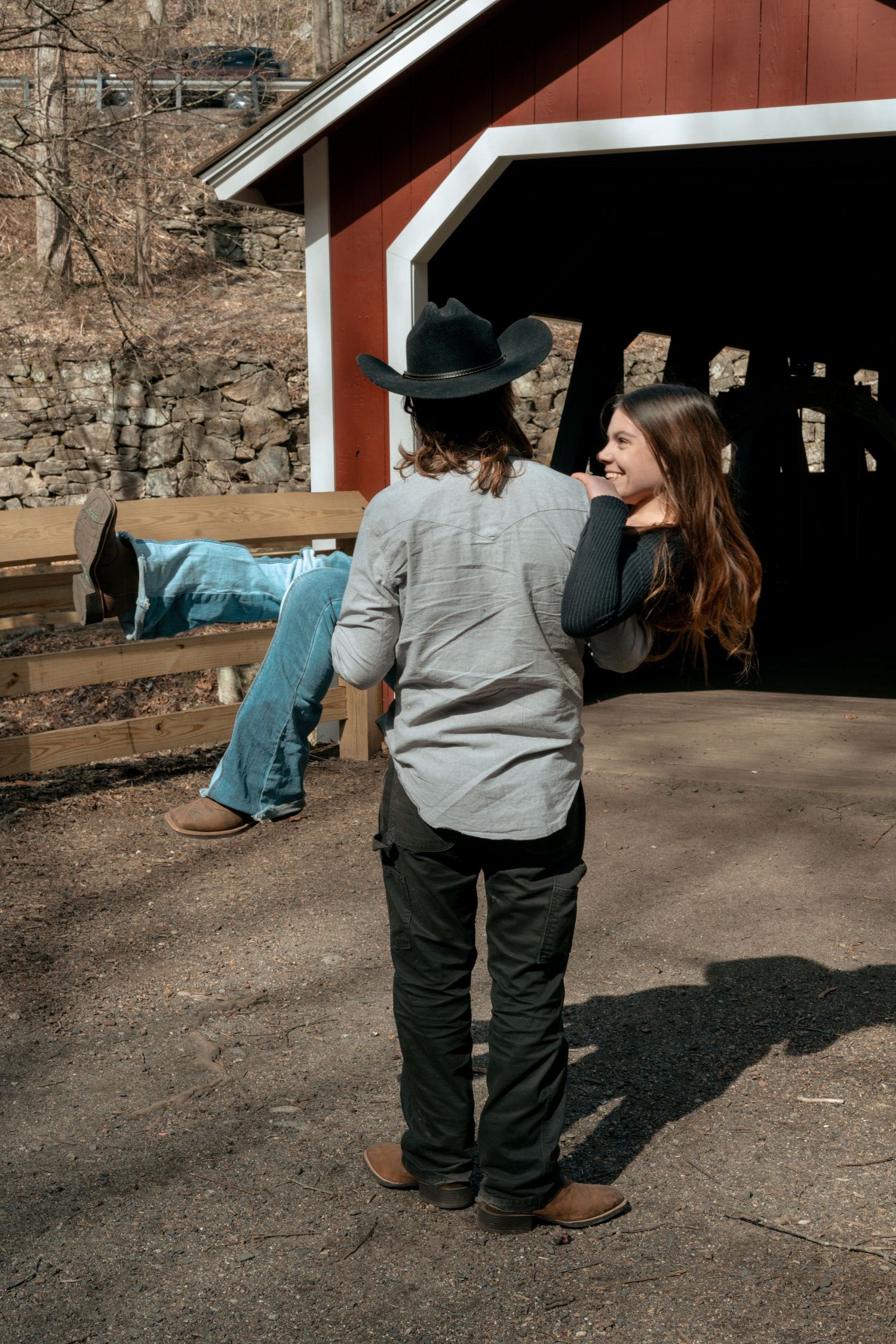 Couple near red covered bridge, woman smiling as man in cowboy hat carries her.