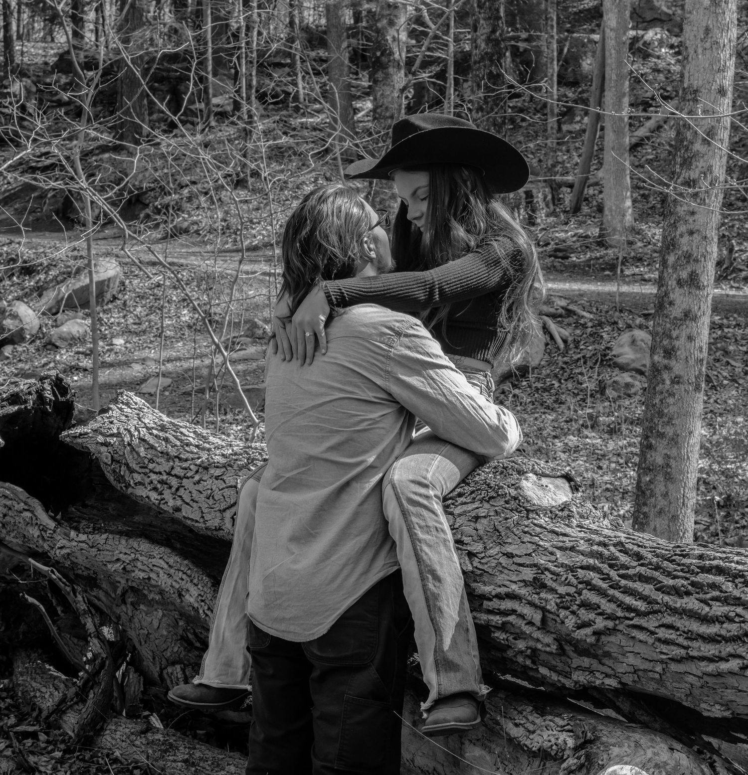 Couple embracing romantically on a fallen log in a forest, black and white engagement photo.