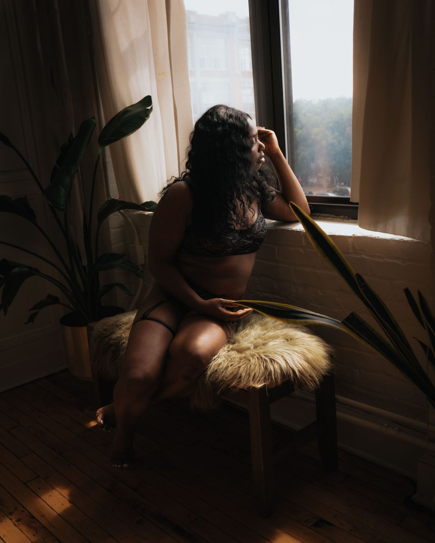 A woman in dark lingerie sits on a fur stool gazing pensively out a sunlit apartment window.