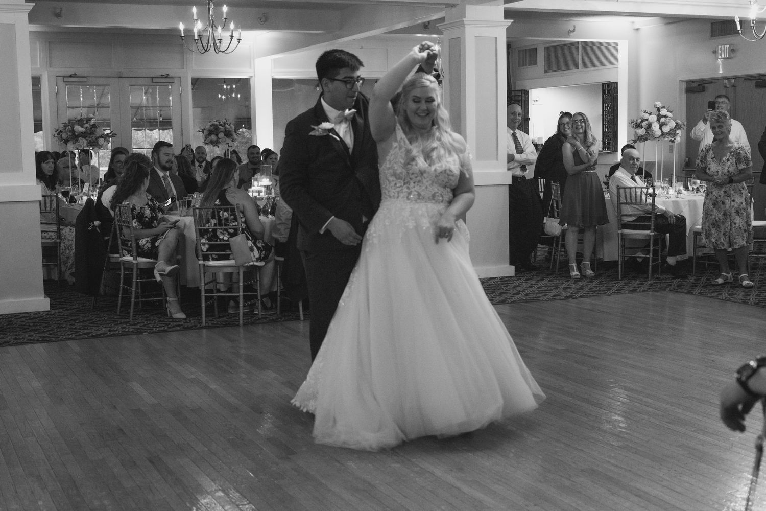 Bride and groom sharing their first dance in a classic ballroom surrounded by wedding guests, black and white.