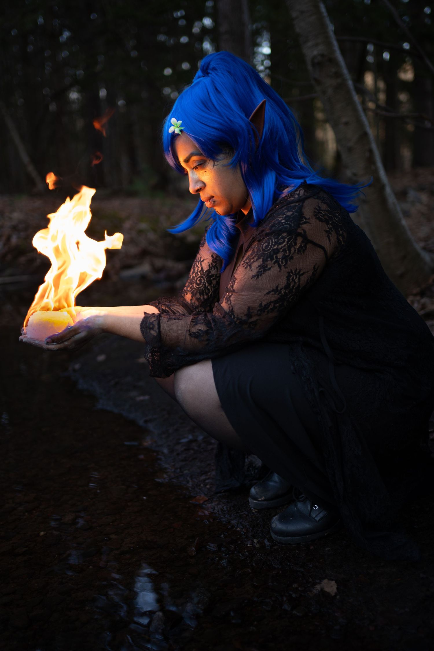 A cosplayer with blue hair and elf ears holds a real flame in her hands while crouching in a dark forest.