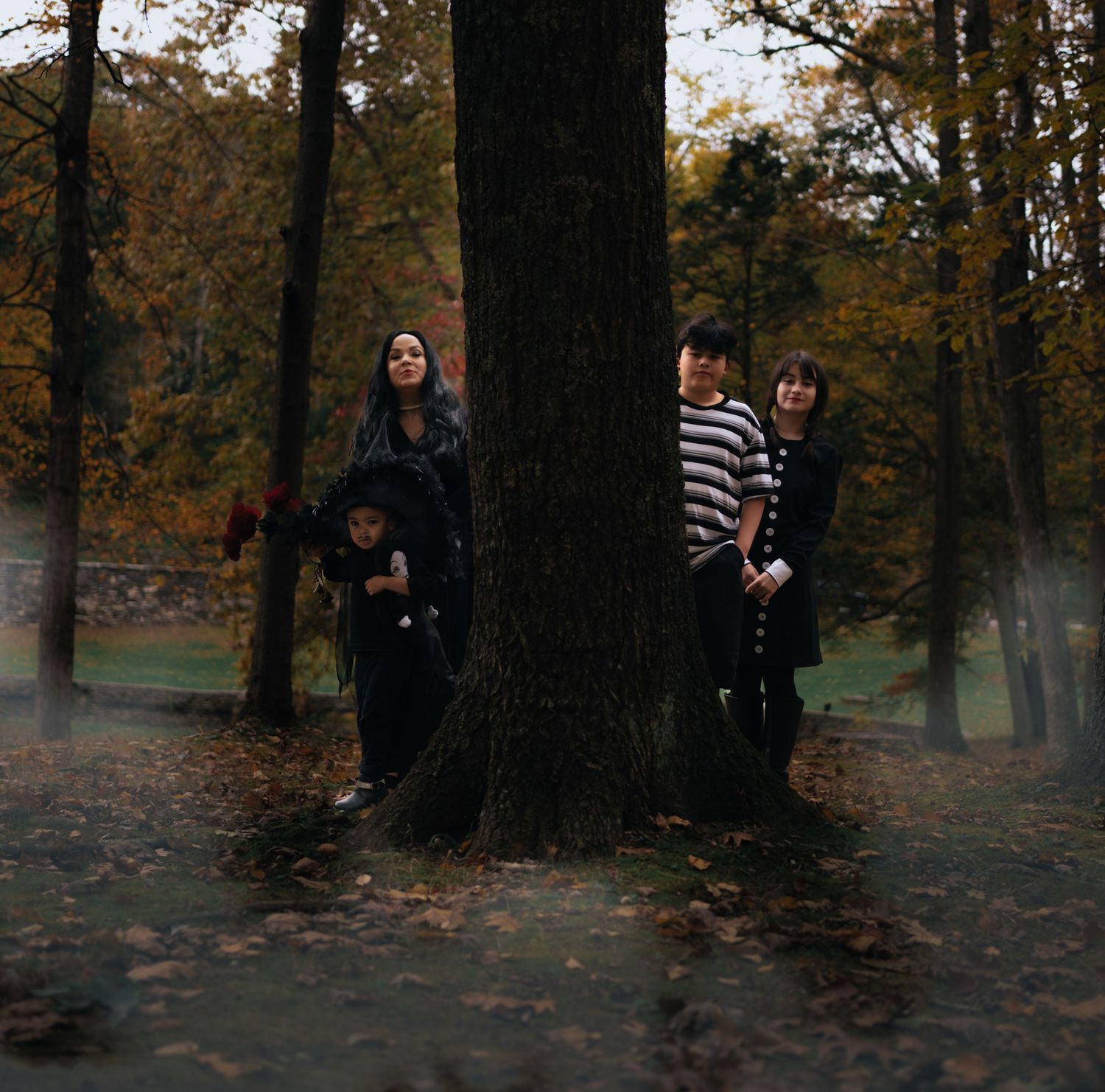 Family of four posing around large tree trunk in misty autumn forest with fallen leaves.