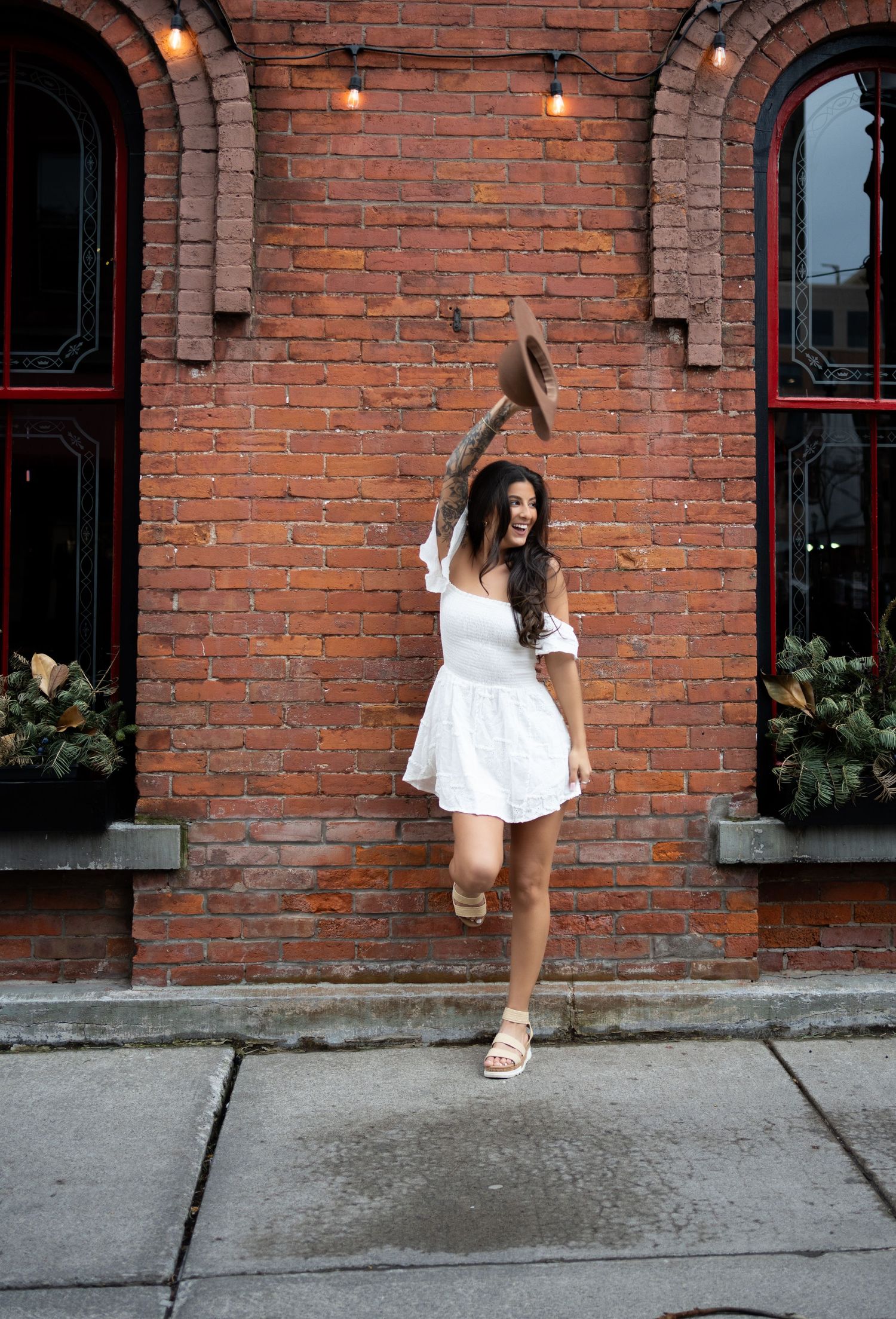 Joyful brunette woman in white mini dress tosses hat in air against rustic brick building.
