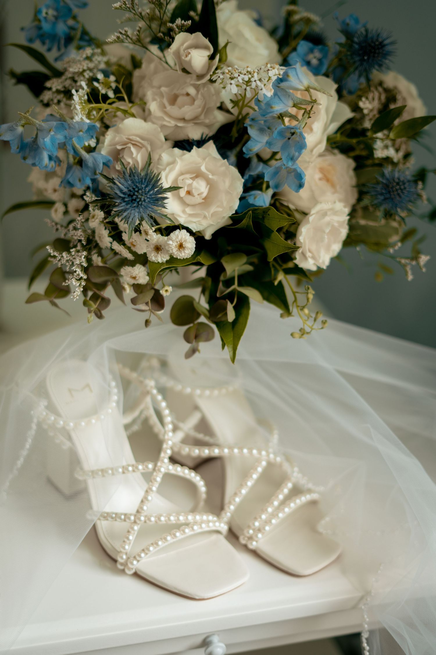 Bridal heels adorned with pearls beside a bouquet of white and blue flowers on white table.