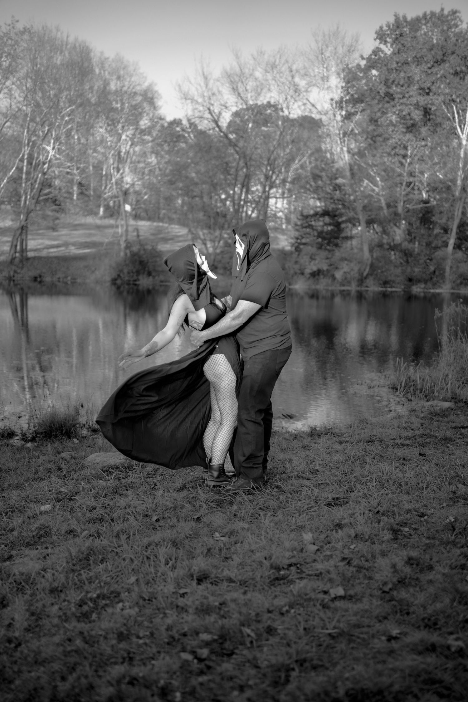 Black and white romantic photo of a couple embracing by a reflective lake surrounded by bare trees.