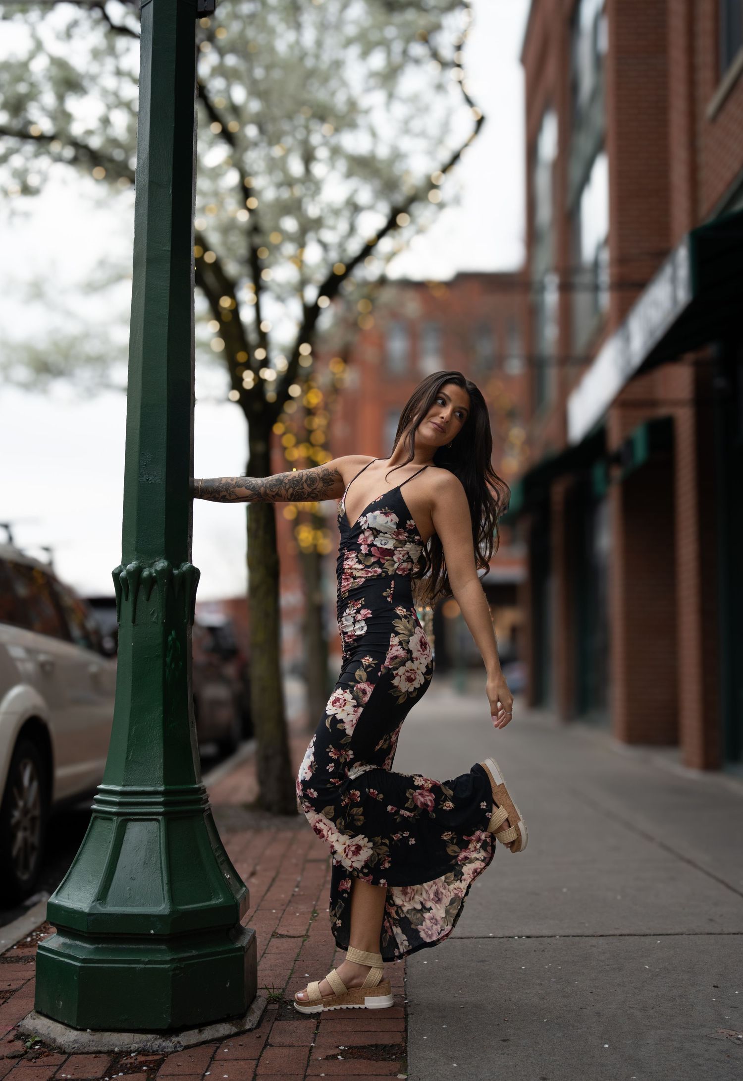 Woman with tattoos twirls in floral maxi dress beside street lamp on charming urban sidewalk.