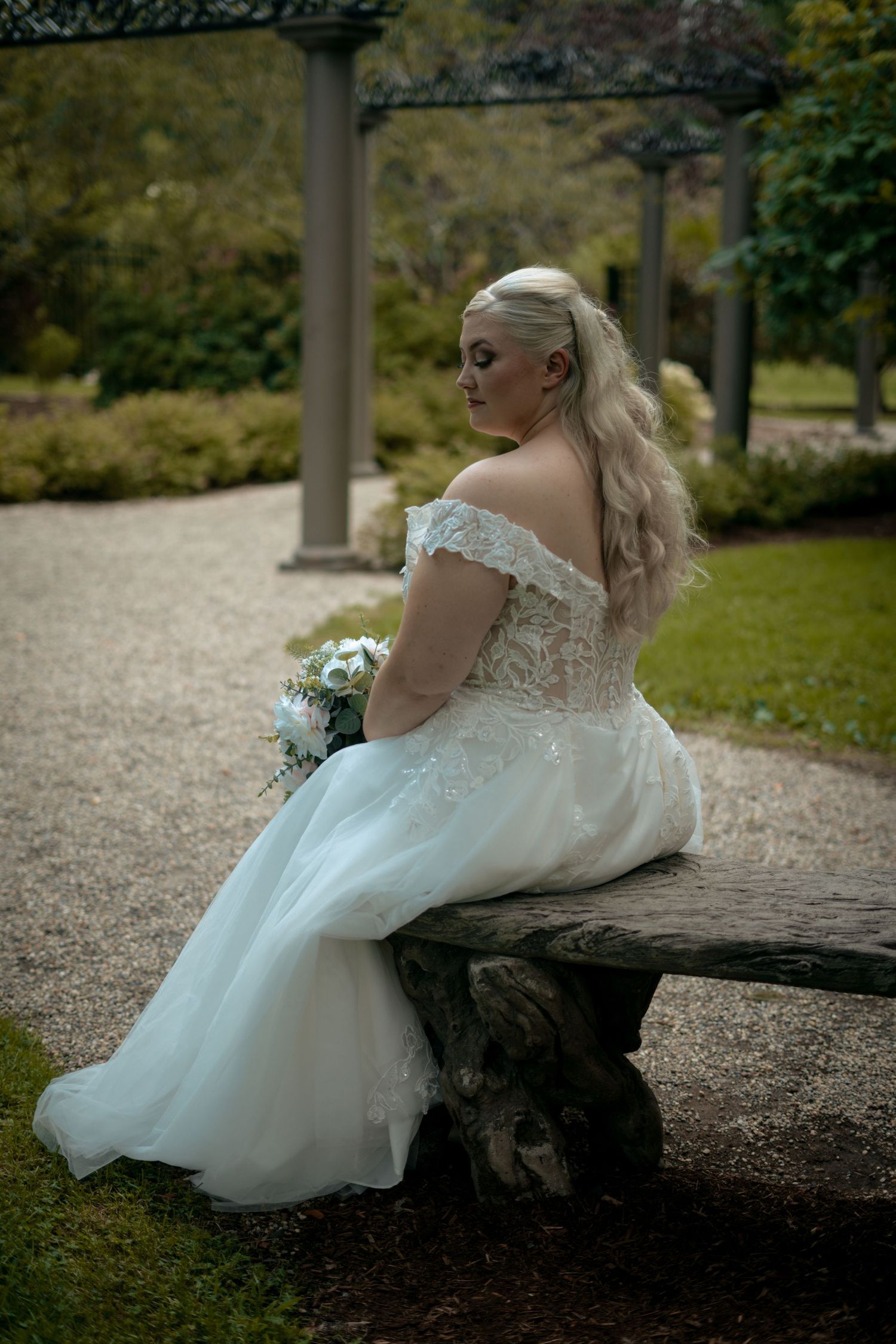 Blonde bride in off-shoulder lace wedding gown sitting on rustic bench in garden.
