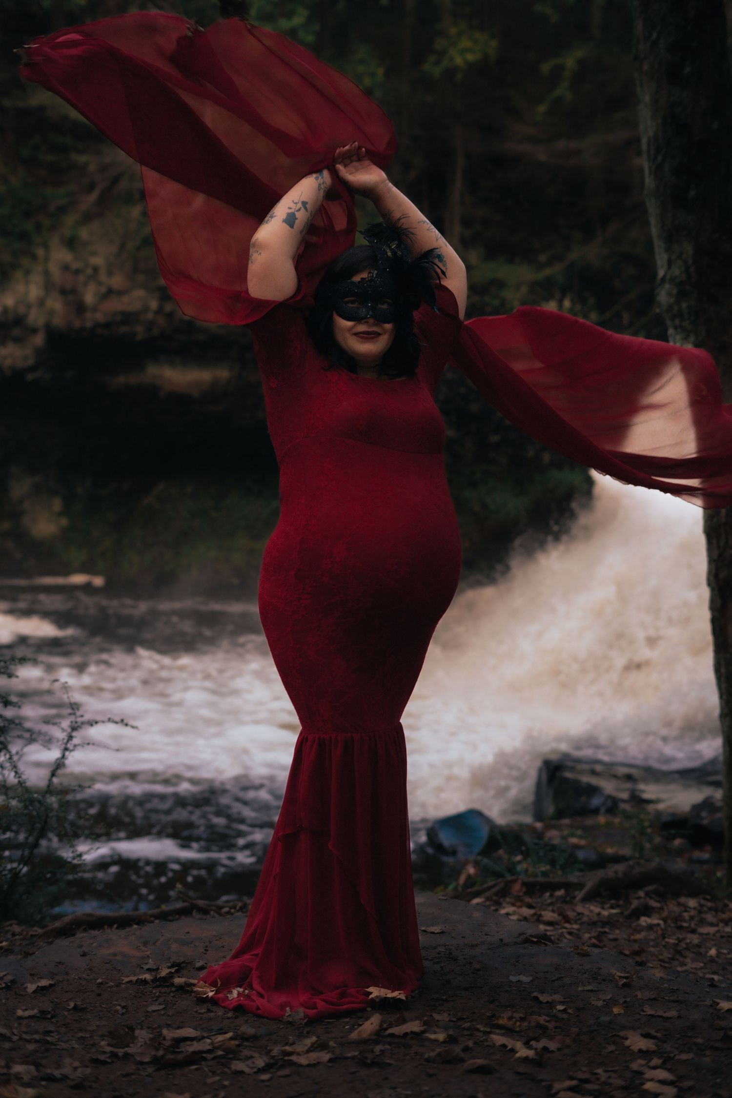 Pregnant woman in red gown wearing black masquerade mask with fabric flowing by waterfall.