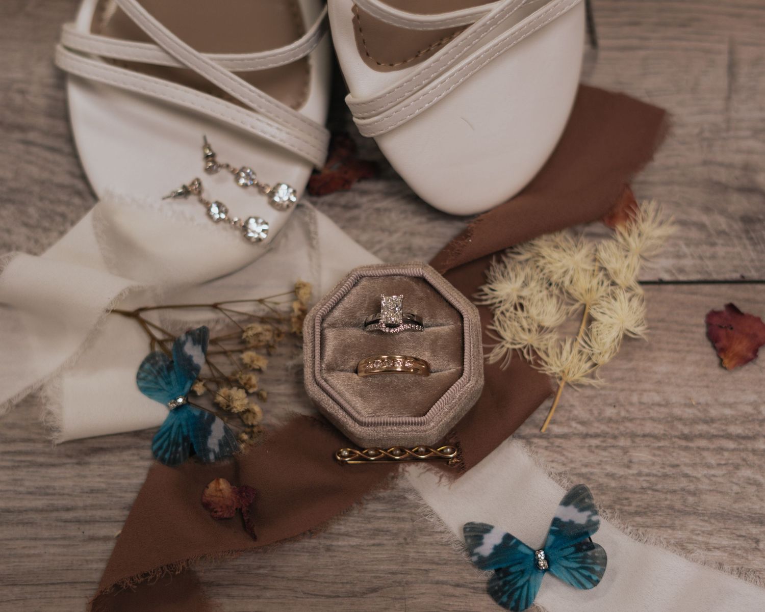 An elegant flat lay of bridal accessories including white shoes, a ring box, earrings, and dried flowers.