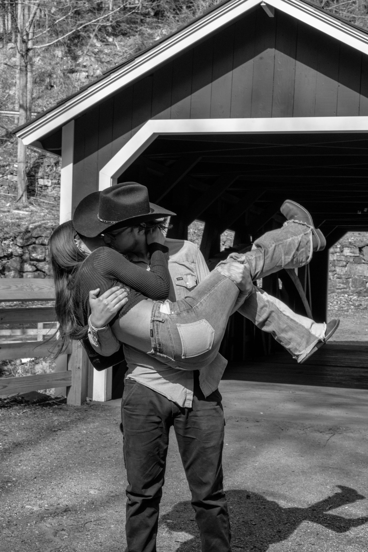Black and white photo of a man in a cowboy hat carrying a woman in his arms near a covered bridge.