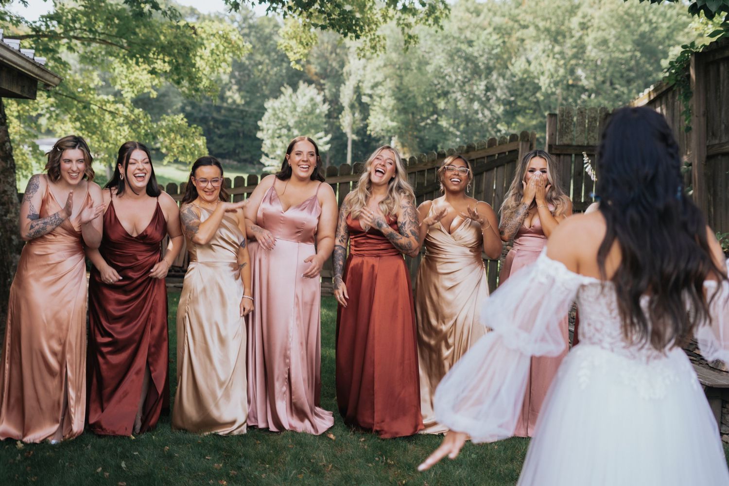 Bridesmaids in satin rose and burgundy gowns reacting with excitement as bride reveals her wedding dress outdoors.