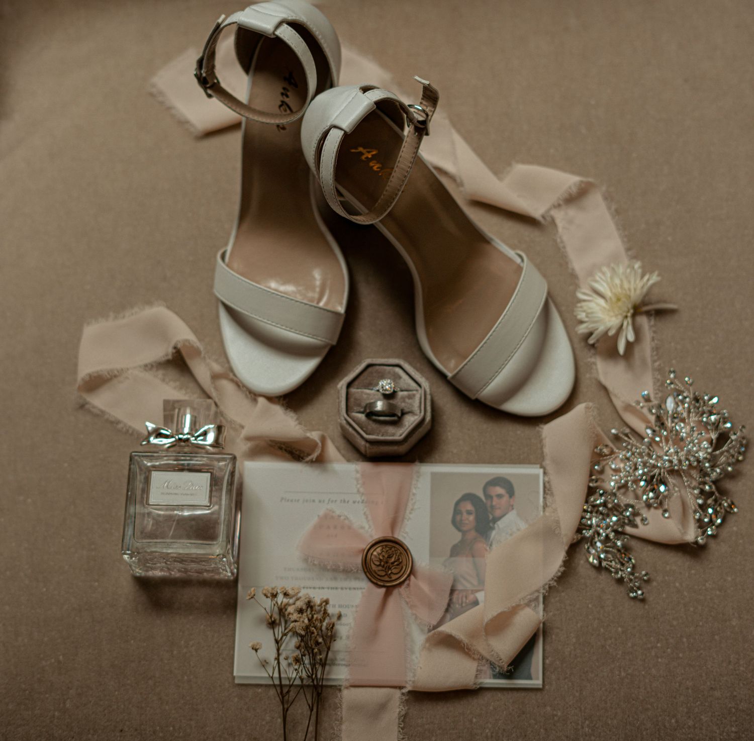 Flat lay of bridal accessories including white heels, perfume, ring box, and a wax-sealed invitation.