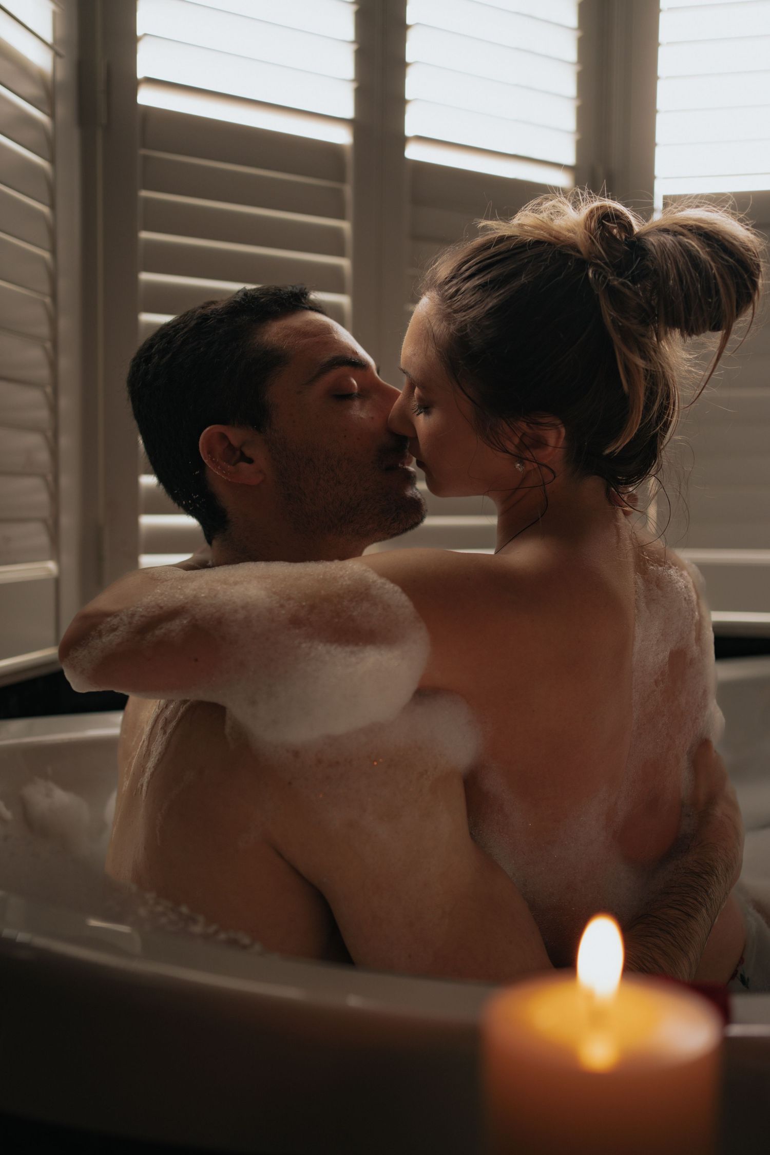 Couple sharing a kiss in a candlelit bubble bath with soft window light.