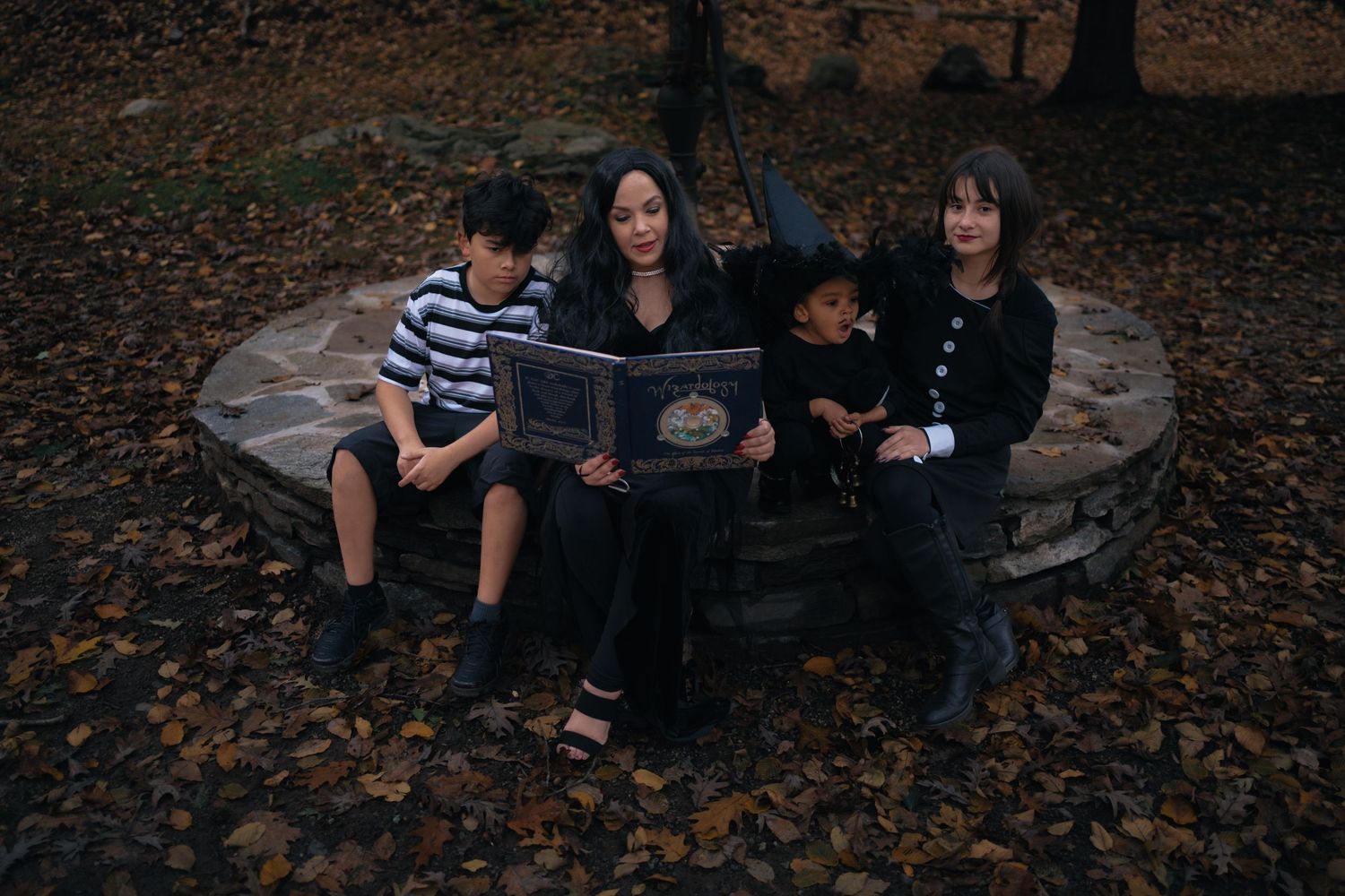 Mother and three children in Halloween costumes reading spooky book on stone bench in autumn.