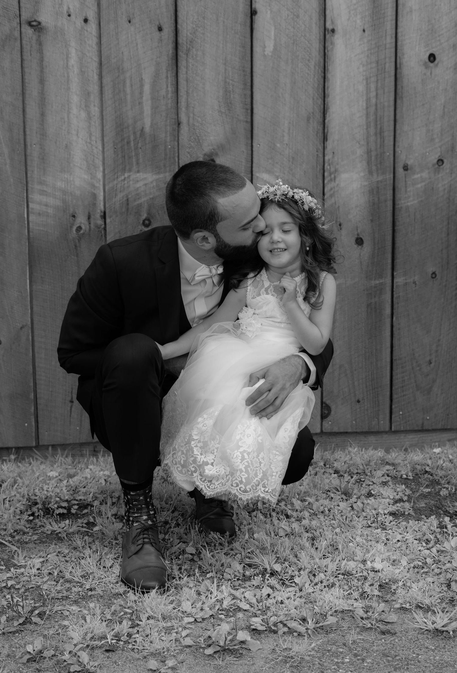 A bearded groom in a suit kisses a young flower girl wearing a white dress and floral crown outdoors.