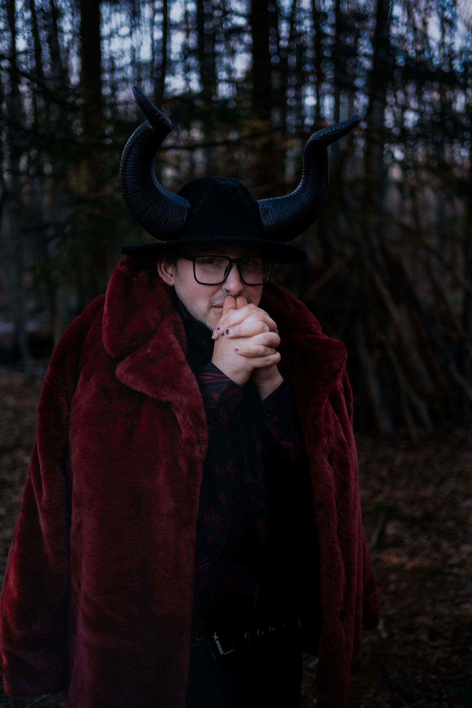 Person wearing large black horns and deep red faux fur coat stands in dark forest setting.