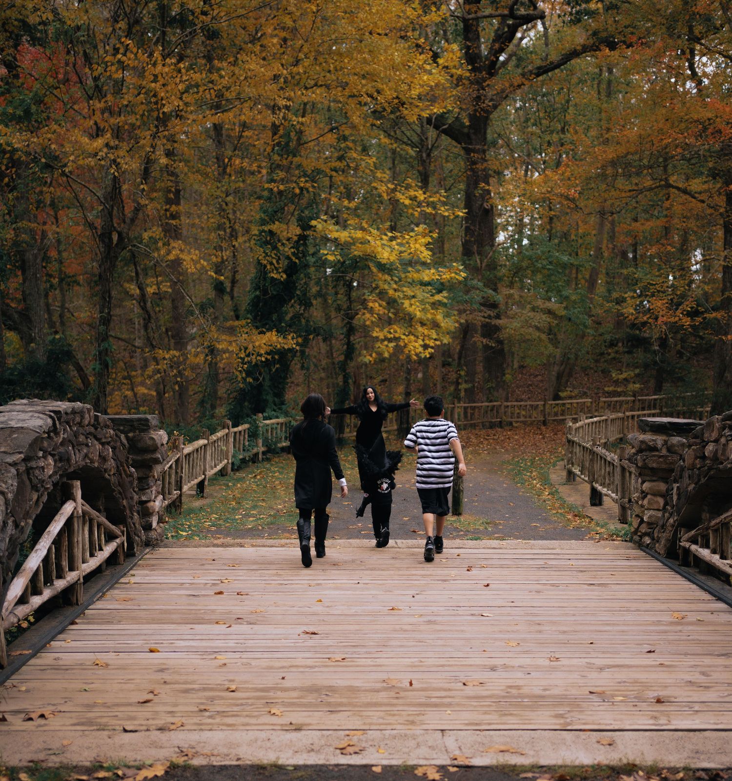 Family in dark costumes walking across wooden bridge surrounded by colorful autumn foliage.