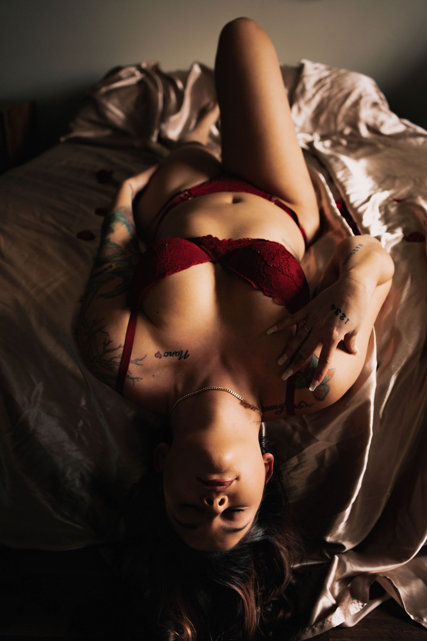 Tattooed woman in red lingerie posing on bed in dramatic moody lighting.