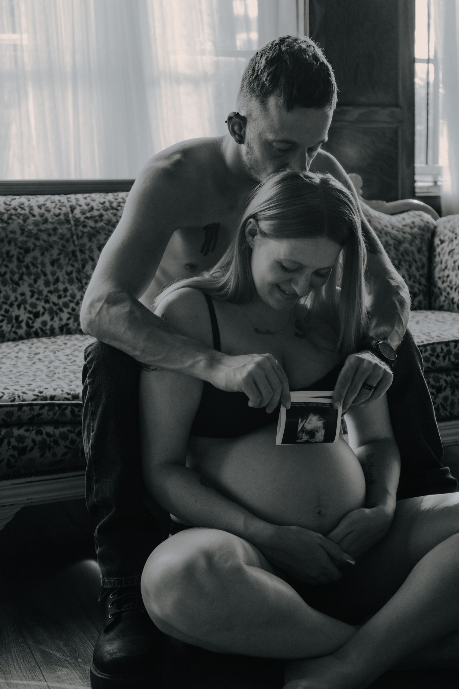 Black and white intimate maternity photo of couple sharing ultrasound moment on vintage floor.