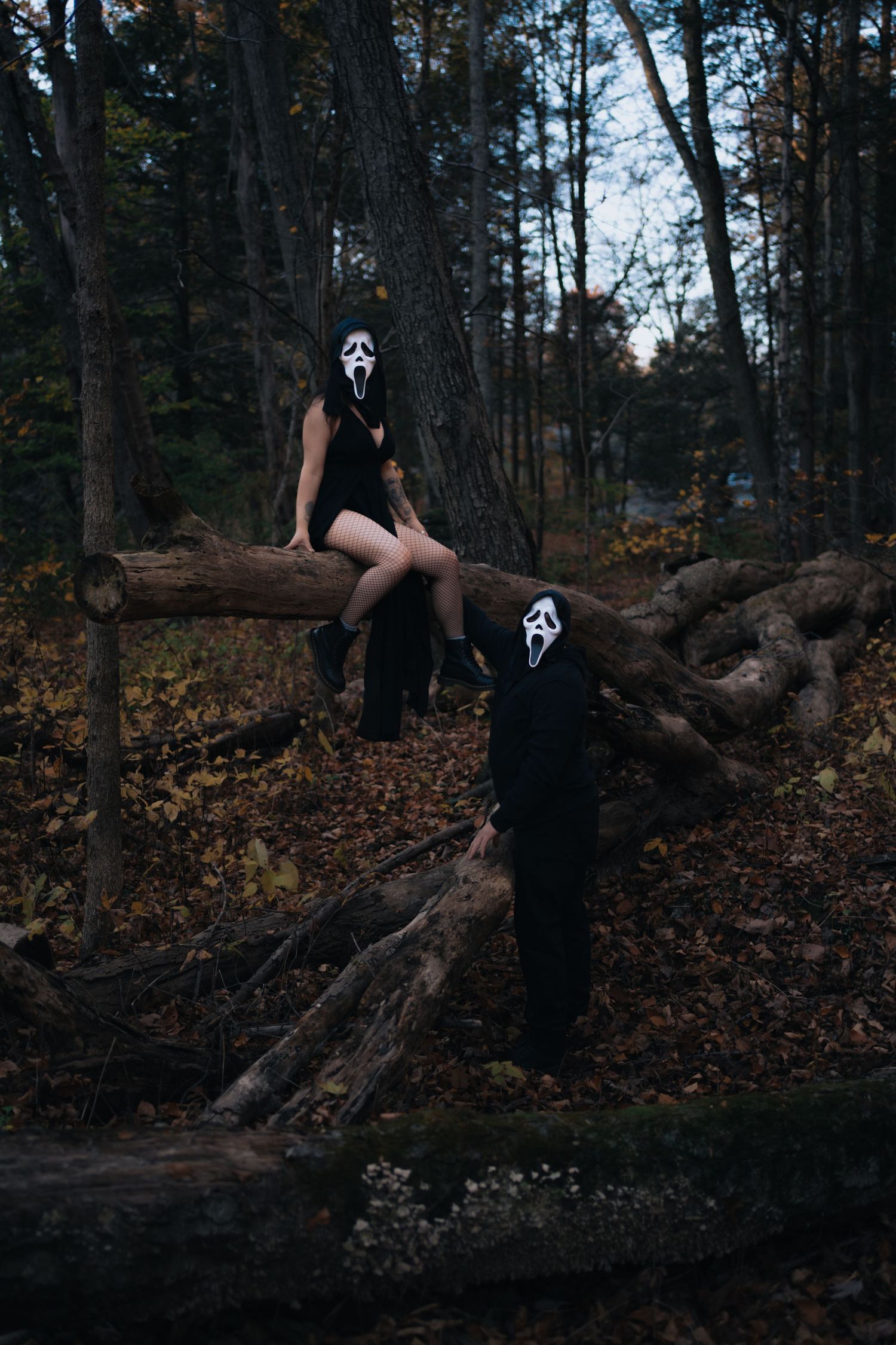 Two people wearing Ghostface Halloween masks posing on fallen logs in an autumn forest at dusk.
