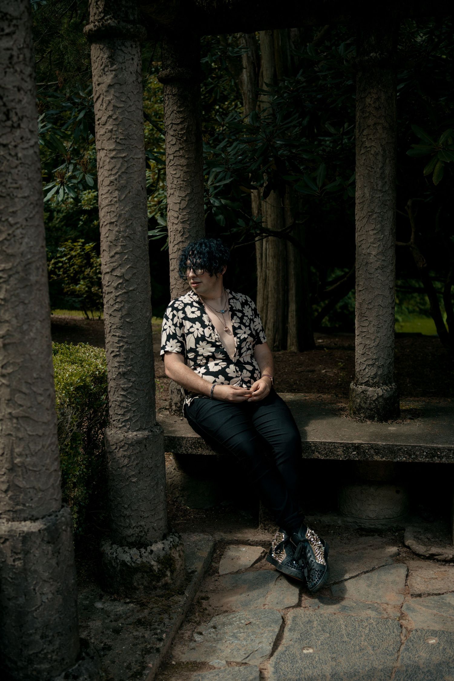 Person in floral shirt sitting between stone columns in shaded garden.