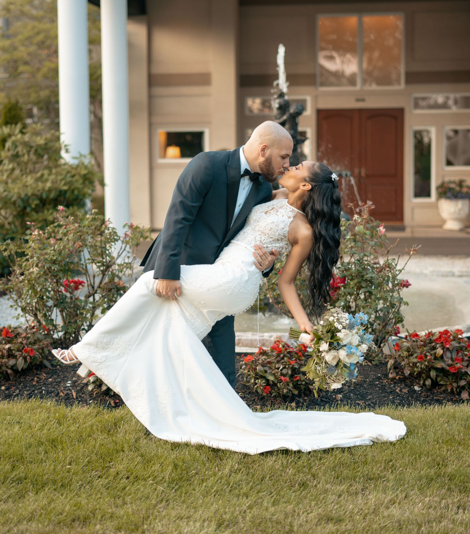Bride and groom sharing a romantic dip kiss in front of a elegant venue surrounded by colorful garden flowers.