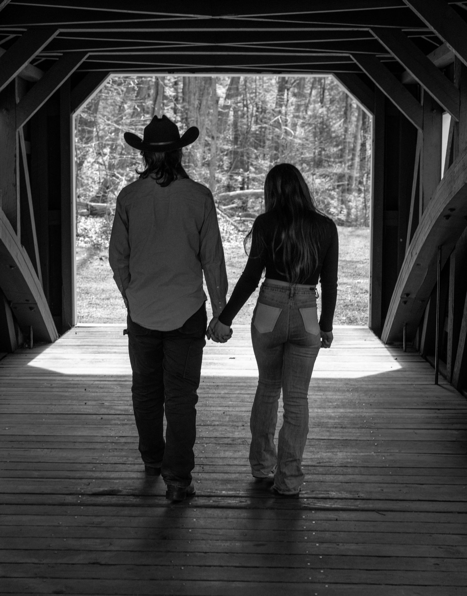 Black and white photo of a couple holding hands walking through a rustic covered wooden bridge surrounded by trees.