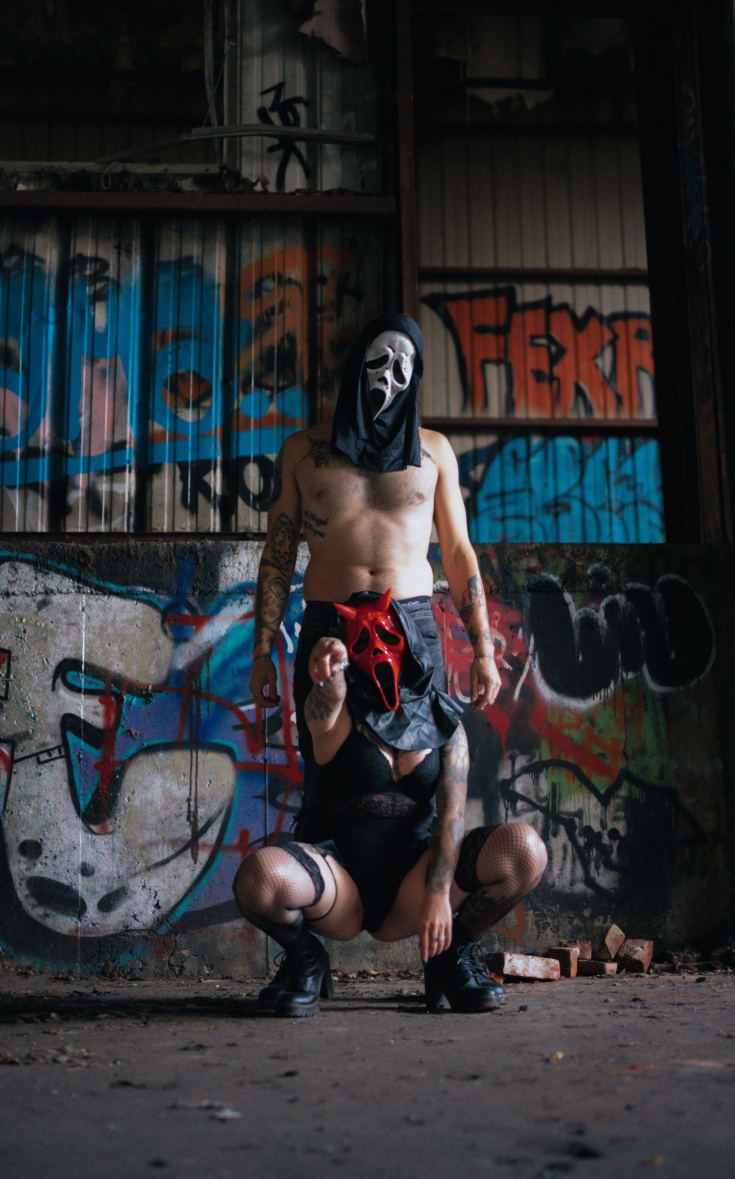 Shirtless tattooed person wearing a Ghostface mask posing in an abandoned building with graffiti walls.