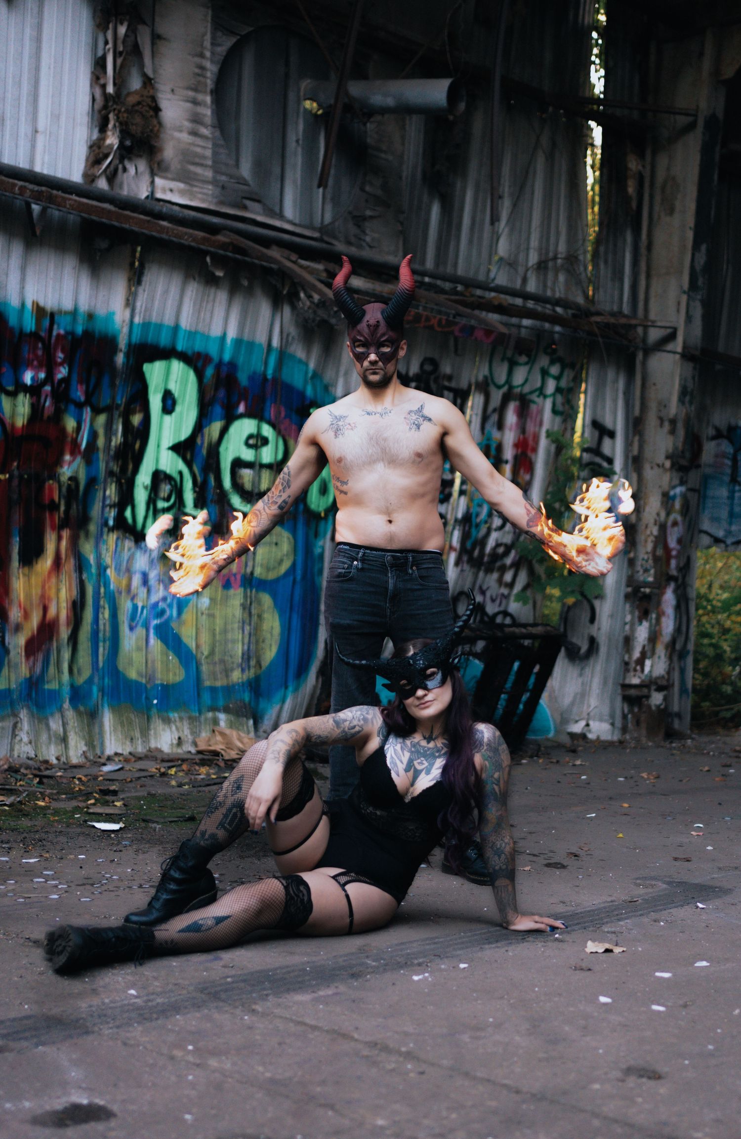 Tattooed couple in devil costumes with fire props pose before graffiti wall in edgy Halloween photoshoot.
