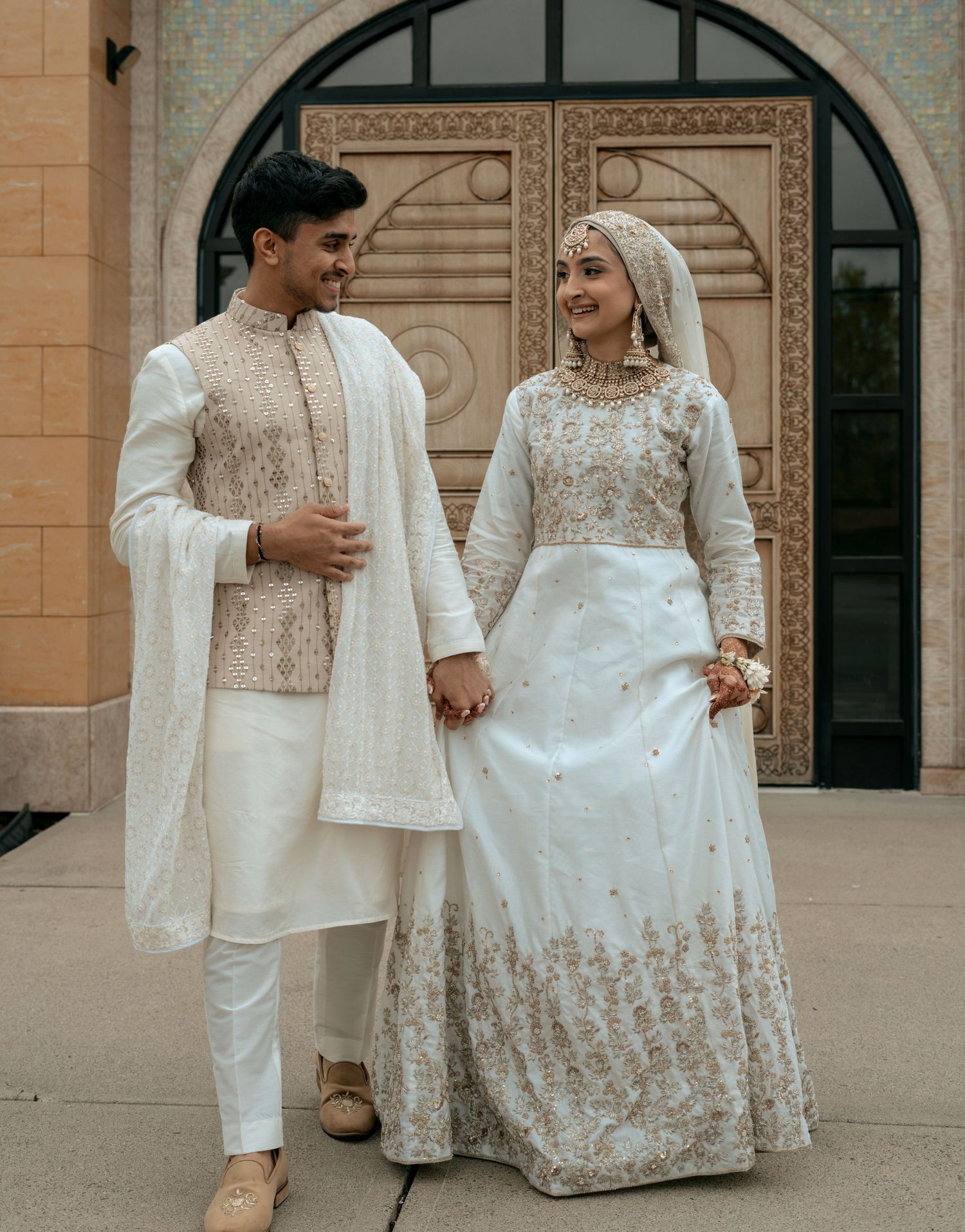 Couple in traditional South Asian wedding attire holding hands and smiling outside an ornate building.