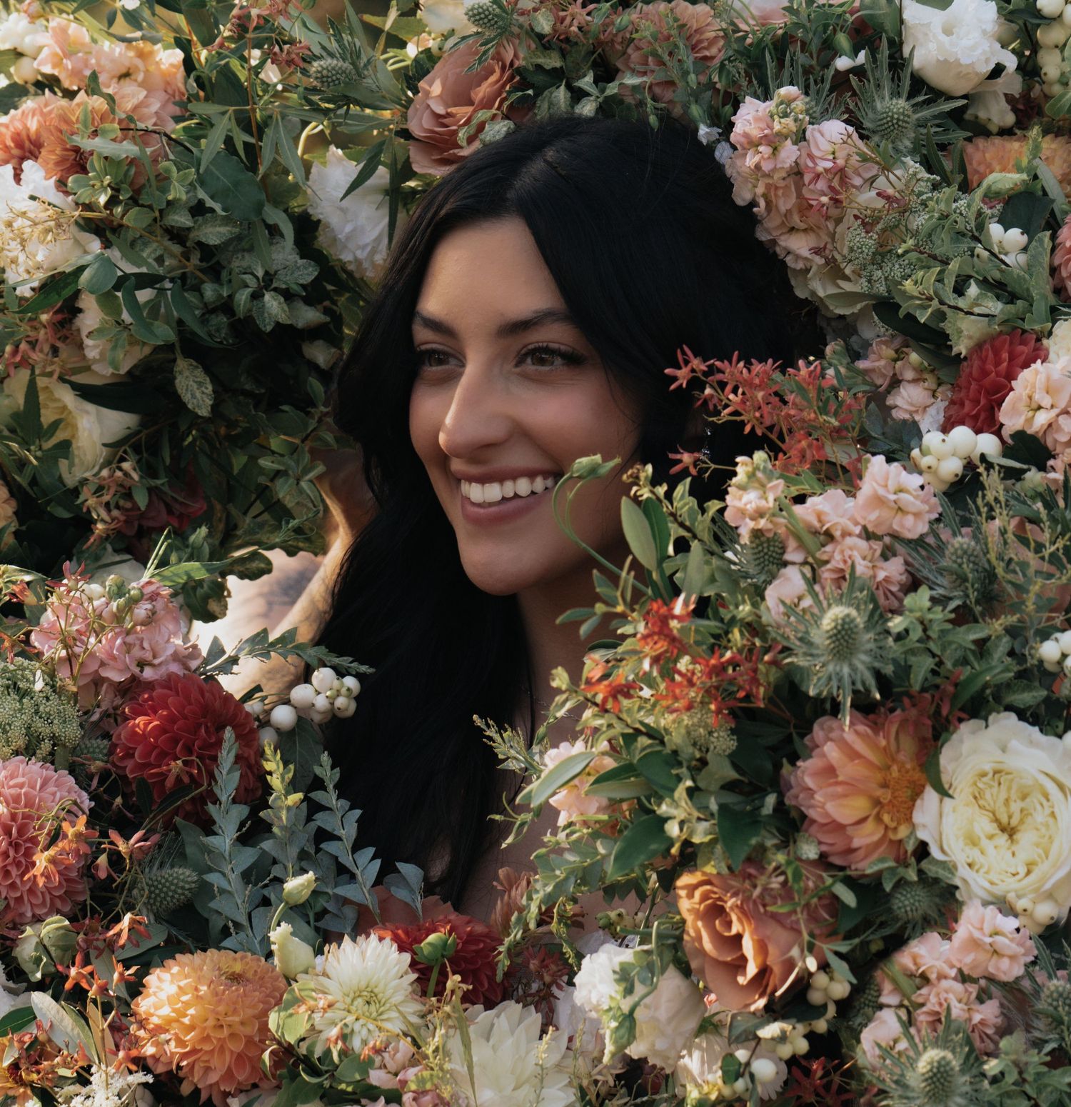 A smiling woman with dark hair peeks through a lush arrangement of colorful flowers including roses and dahlias.