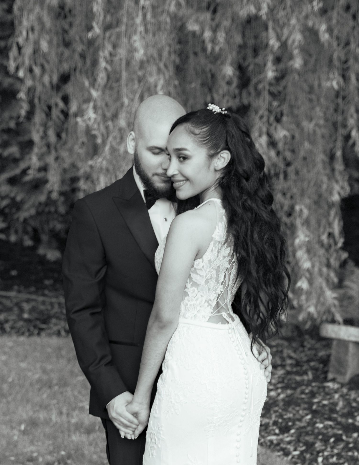 A black and white wedding portrait of a bride and groom embracing beneath a weeping willow tree.