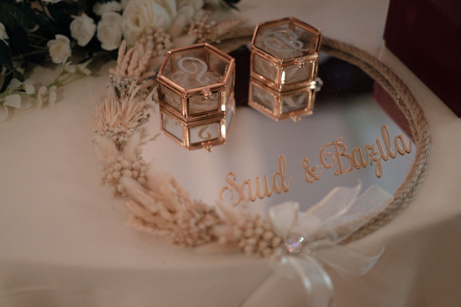 Gold hexagonal ring boxes and a dried floral wreath on a table engraved with the names Saud and Bazila.